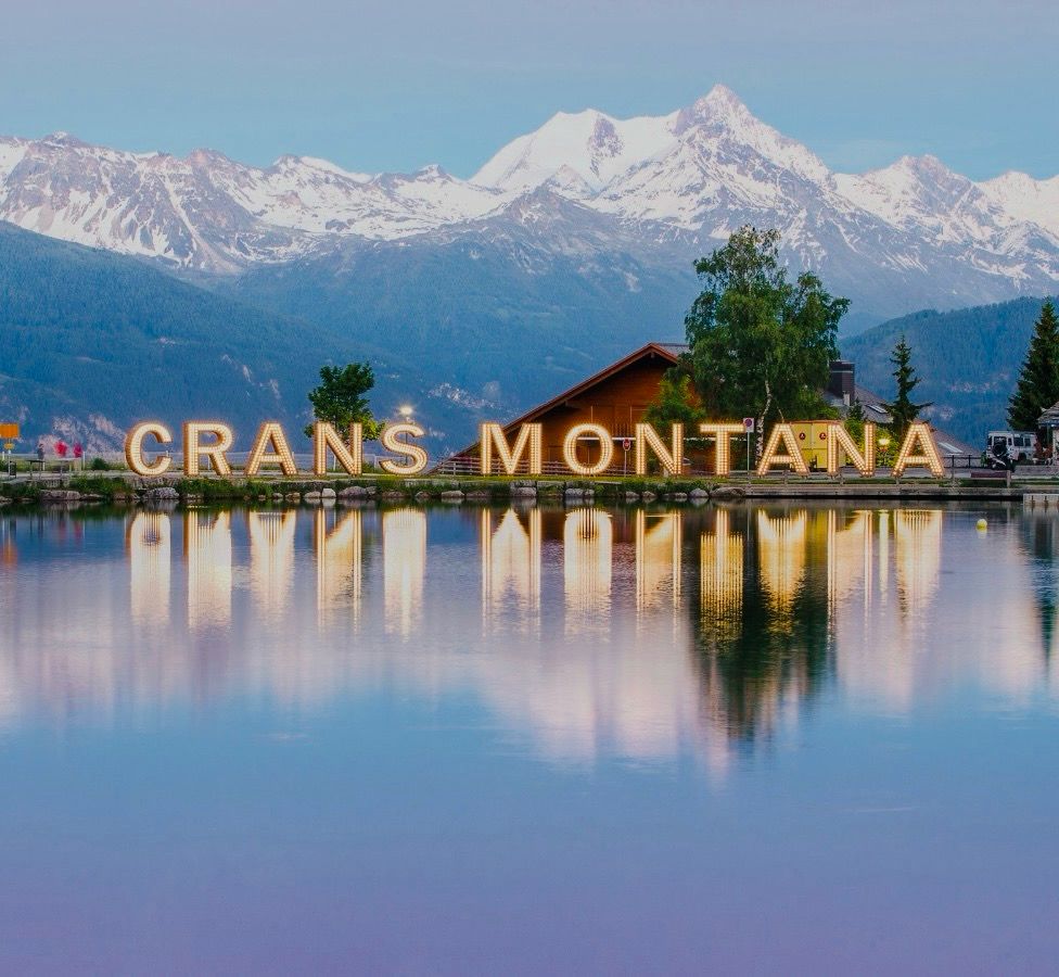 Crans-montana-lac-valais.Lac calme avec reflets des montagnes enneigées et grandes lettres lumineuses formant 'Crans Montana', avec un chalet en bois et des arbres au bord du lac sous un ciel clair.